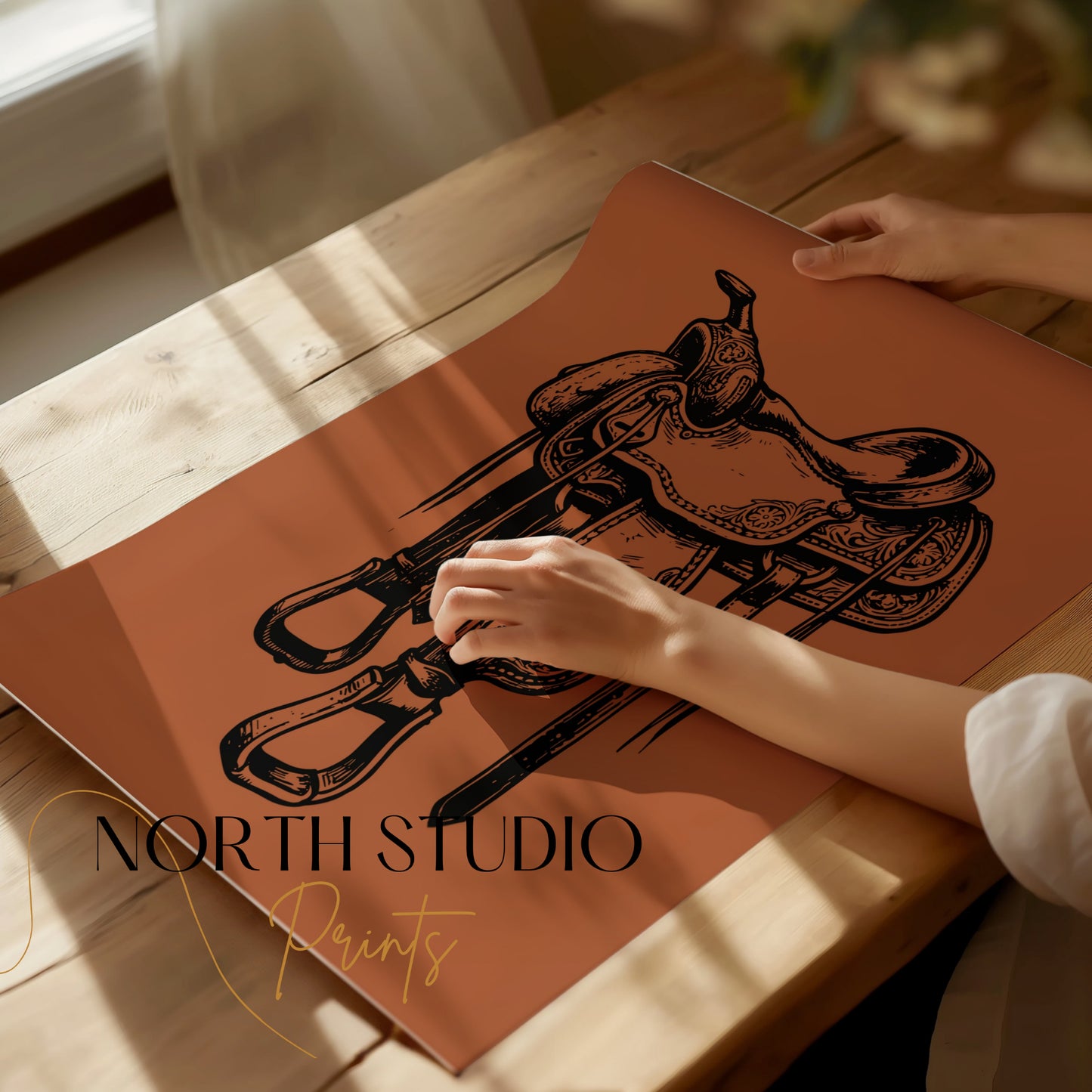 Western saddle illustration on copper paper, hands holding edge, "North Studio Prints" logo.