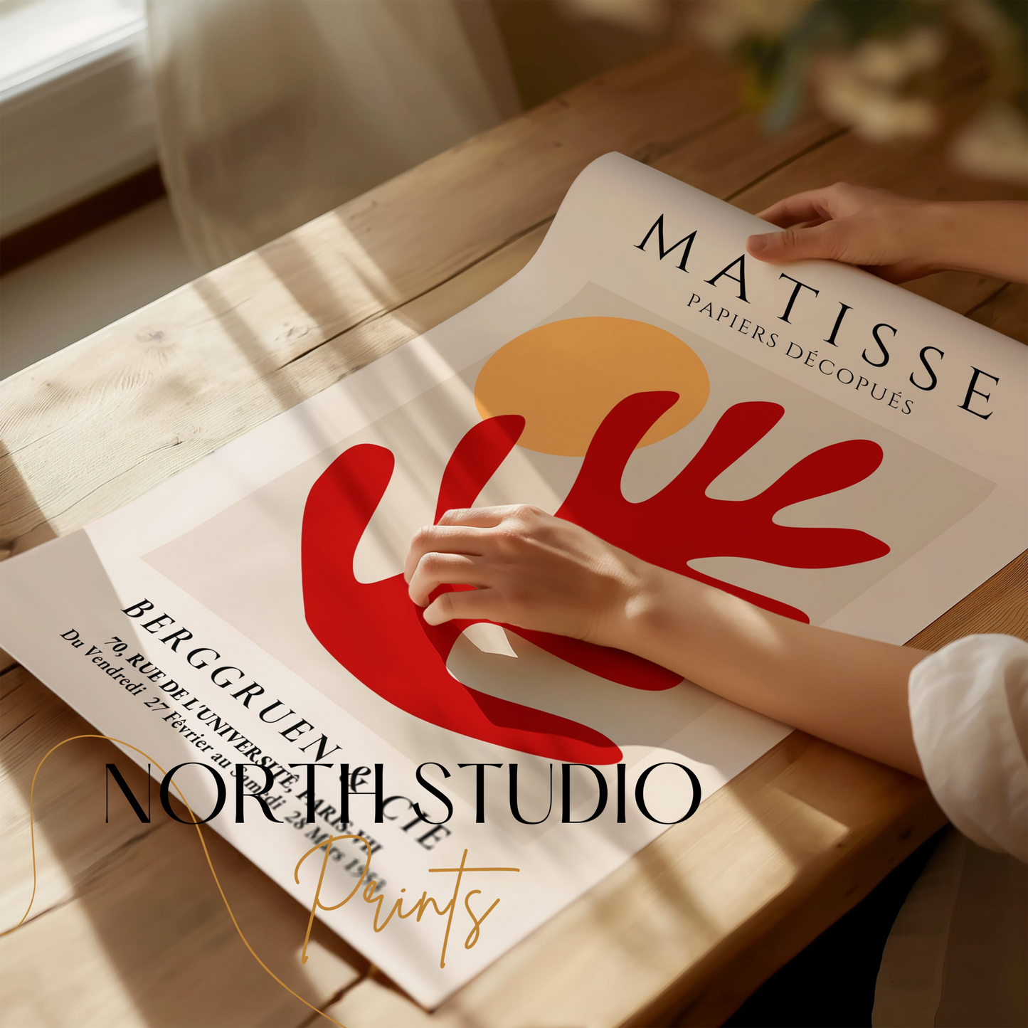 Hands holding an unframed abstract red leaf and sun Matisse art exhibition poster on a wooden table, emphasizing its minimalist and modern design.