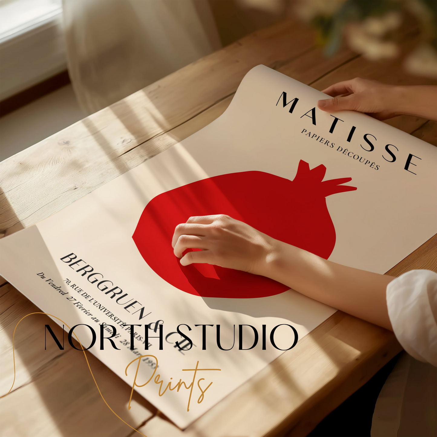 Hands unrolling a Matisse Papiers Decoupes Berggruen & Cie poster with a red pomegranate design on a wooden table, branded with North Studio Prints.