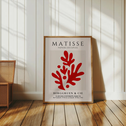 Framed Matisse Papiers Découpés poster with red abstract cutout design, displayed leaning against a white wall in a sunlit room with wooden flooring.