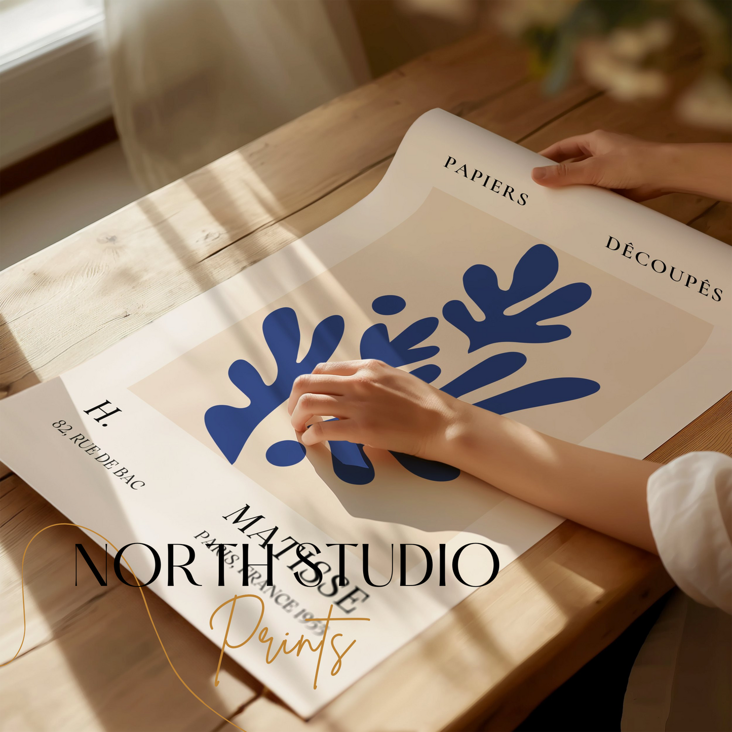Close-up of a Matisse-inspired decorative poster with blue leaf cut-outs, being unrolled on a wooden table with soft natural light.