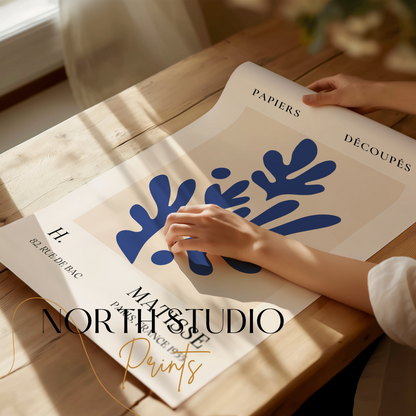 Close-up of a Matisse-inspired decorative poster with blue leaf cut-outs, being unrolled on a wooden table with soft natural light.