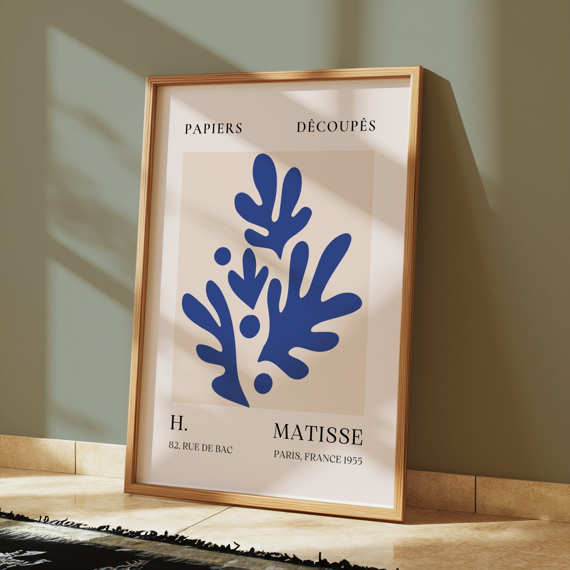 Abstract blue leaf cut-out poster in a wooden frame, styled on a tiled floor with sunlight streaming through a nearby window.