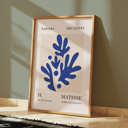 Abstract blue leaf cut-out poster in a wooden frame, styled on a tiled floor with sunlight streaming through a nearby window.