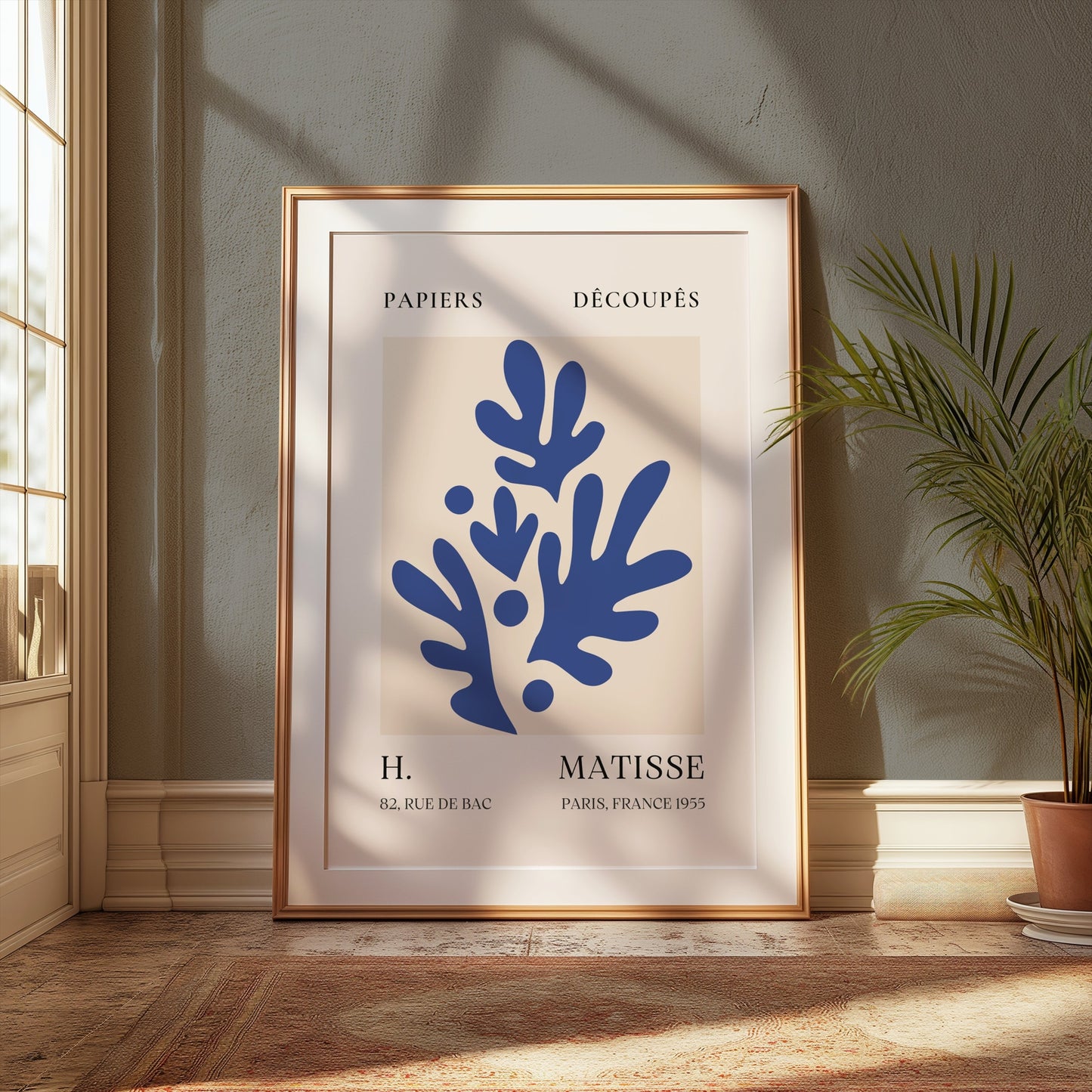 Matisse-inspired decorative poster with blue leaf cut-outs, framed and styled in a cozy room with a potted plant and warm lighting.