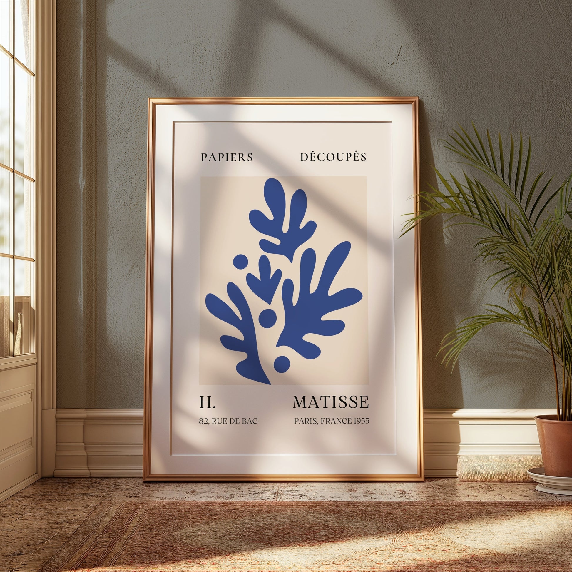 Matisse-inspired decorative poster with blue leaf cut-outs, framed and styled in a cozy room with a potted plant and warm lighting.