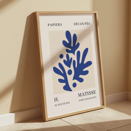 Elegant Parisian art print in Matisse style with blue botanical shapes, framed and leaning against a beige wall in a sunlit room.