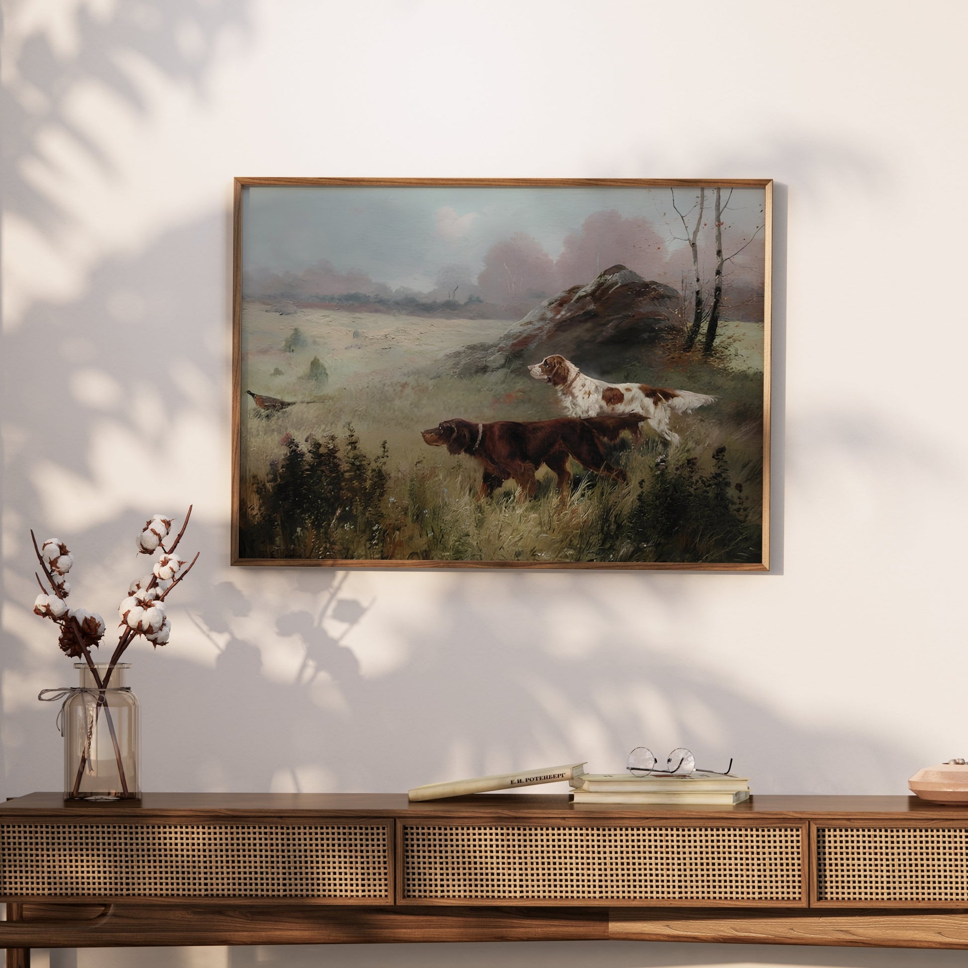 Framed hunting dogs in misty forest landscape poster styled on a console table with books and minimalist decor.