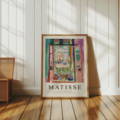 Matisse The Open Window 1905 poster in a wooden frame, leaning against a white paneled wall with natural light and wicker furniture.