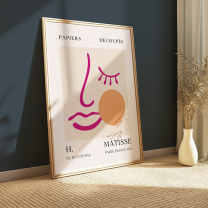 Modern abstract face art inspired by Matisse posters in a gold frame, leaning against a dark teal wall with minimalist decor.