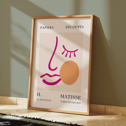 Abstract face art inspired by Matisse posters in a wooden frame, displayed on a tiled floor with soft sunlight streaming in.