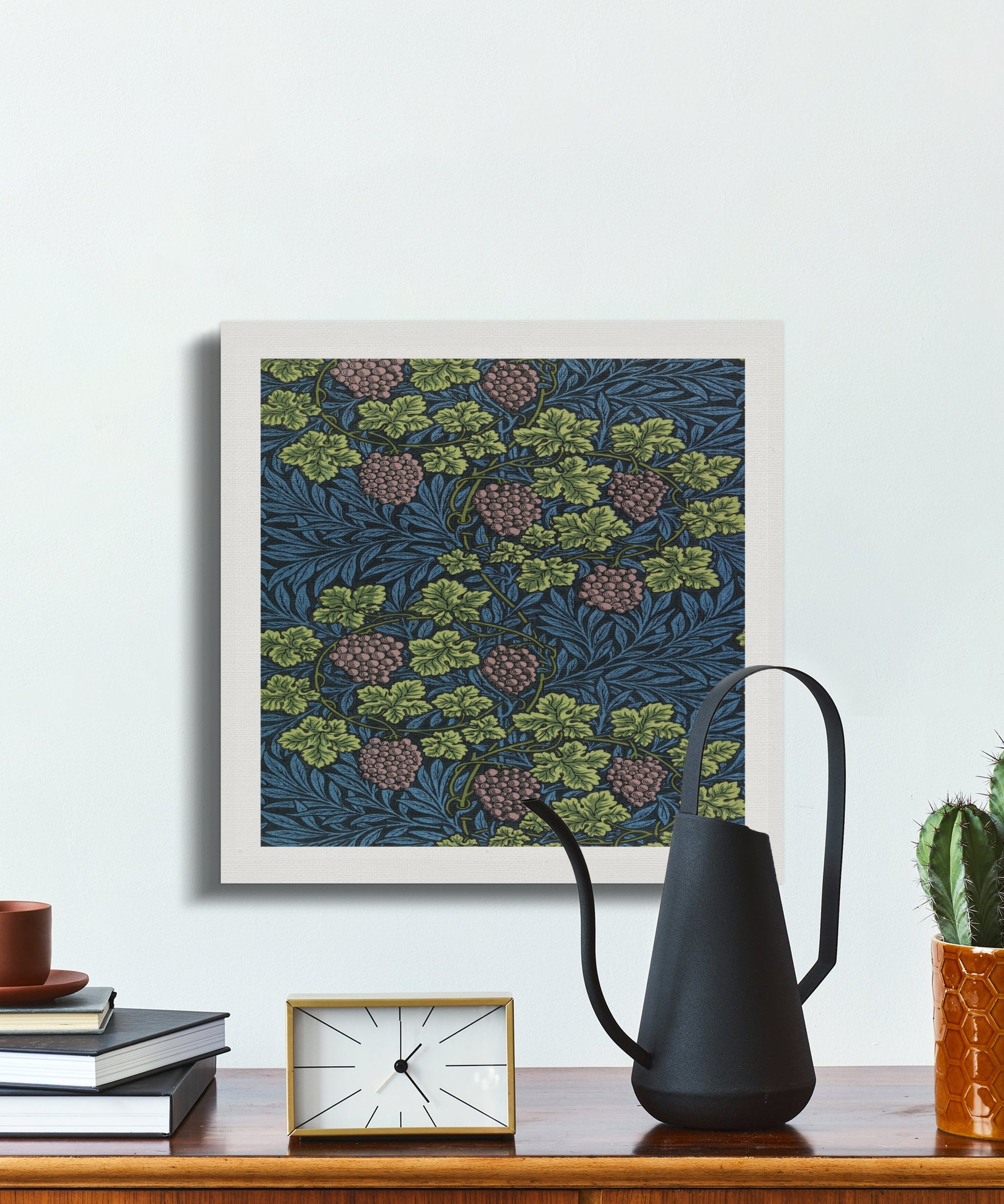 William Morris Grape Vines canvas wall art displayed in a modern interior with a clock, watering can, and potted plants on a wooden surface.